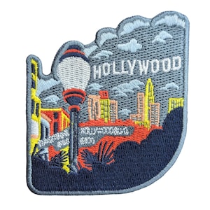 May include: Embroidered patch depicting Hollywood. The design includes a street lamp, cityscape, and the word "HOLLYWOOD" in white lettering against a blue sky. The patch has a dark blue border, suitable for application on clothing or bags.