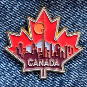 Può includere: Una spilla in smalto rosso e oro a forma di foglia d'acero. La spilla presenta uno skyline con la CN Tower e la scritta "CANADA" in lettere bianche. La spilla è ambientata su uno sfondo di denim.