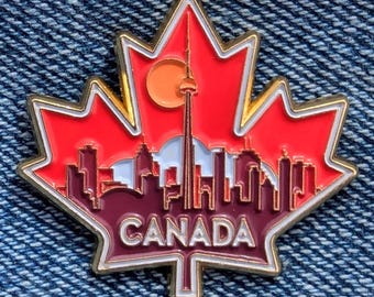 May include: A red and gold enamel pin in the shape of a maple leaf. The pin features a skyline with the CN Tower and the word "CANADA" in white lettering. The pin is set against a denim background.