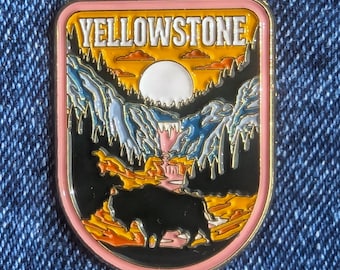 May include: A rectangular enamel pin with a pink border featuring the text "YELLOWSTONE". The pin depicts a landscape with mountains, a waterfall, a bison, and a sunset in shades of yellow, orange, blue, and black.