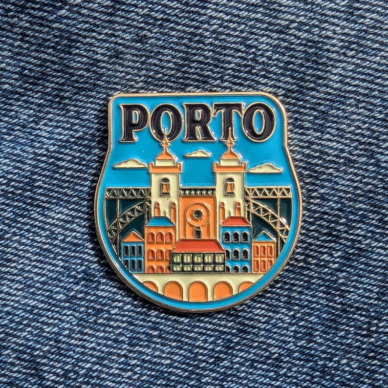 May include: A gold-colored enamel pin with a blue background and a white outline. The pin features a stylized image of the city of Porto, Portugal, with the text "PORTO" in black letters at the top. The image includes buildings, a bridge, and clouds.