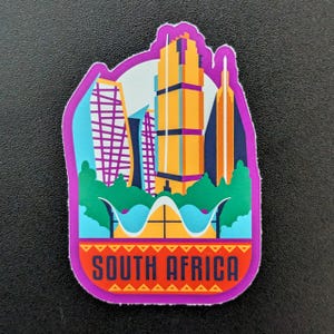 Op de afbeelding: Een kleurrijke sticker met een stadsgezicht met hoge gebouwen in oranje, paars en blauw. De sticker heeft een paarse rand en de woorden "SOUTH AFRICA" in rood en oranje. De achtergrond is lichtblauw.