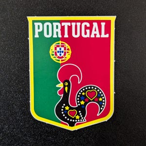 May include: A rectangular sticker with a yellow border and the word "PORTUGAL" in white. The sticker features the Portuguese flag colors, a rooster design, and a coat of arms.