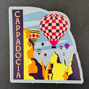May include: A sticker featuring a colorful illustration of hot air balloons over a landscape. The sticker has the word "Cappadocia" in vertical text. The balloons are in various colors, including pink, yellow, and blue.