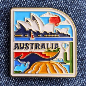 May include: A rectangular enamel pin depicting Australian landmarks. The pin showcases the Sydney Opera House, a red sun, a kangaroo, and the word "AUSTRALIA". The design incorporates blue, white, red, yellow, and green colours.