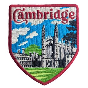 Könnte beinhalten: Ein schildförmiger gestickter Aufnäher mit dem Wort Cambridge in roter Schrift am oberen Rand. Das Design zeigt ein Steingebäude mit zwei hohen Türmen, einen hellblauen Himmel mit weißen Wolken, grüne Bäume und eine grüne Wiese, alles rot umrandet.