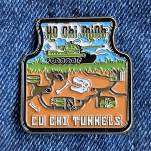 May include: A rectangular pin featuring a green tank and the text "Ho Chi Minh" above a depiction of the Cu Chi Tunnels. The pin has a black border and a blue, green, and orange color scheme. The text "Cu Chi Tunnels" is at the bottom.