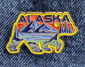 May include: A colorful Alaska enamel pin in the shape of a bear. The pin features the word "ALASKA" in yellow letters above a landscape scene with mountains, a lake, and trees. The pin has a yellow border and is set against a denim background.