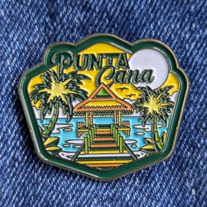 May include: A colorful enamel pin featuring a tropical beach scene with the words "Punta Cana". The design includes palm trees, a pier, and a gazebo, set against a yellow and blue background, framed by a green border.