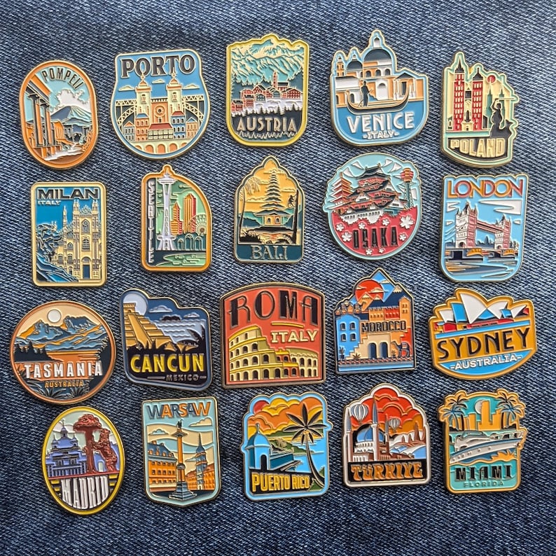 May include: A collection of 20 enamel pins featuring various cityscapes and landmarks from around the world. The pins are colorful and detailed, with gold accents. The pins are arranged in a grid pattern on a blue denim background.