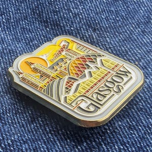 Peut inclure: &Eacute;pinglette rectangulaire repr&eacute;sentant un paysage urbain stylis&eacute; de Glasgow, avec le mot "Glasgow" en police d&eacute;corative. La broche a une bordure dor&eacute;e et un design aux tons jaunes, rouges et gris, sur fond de denim bleu.