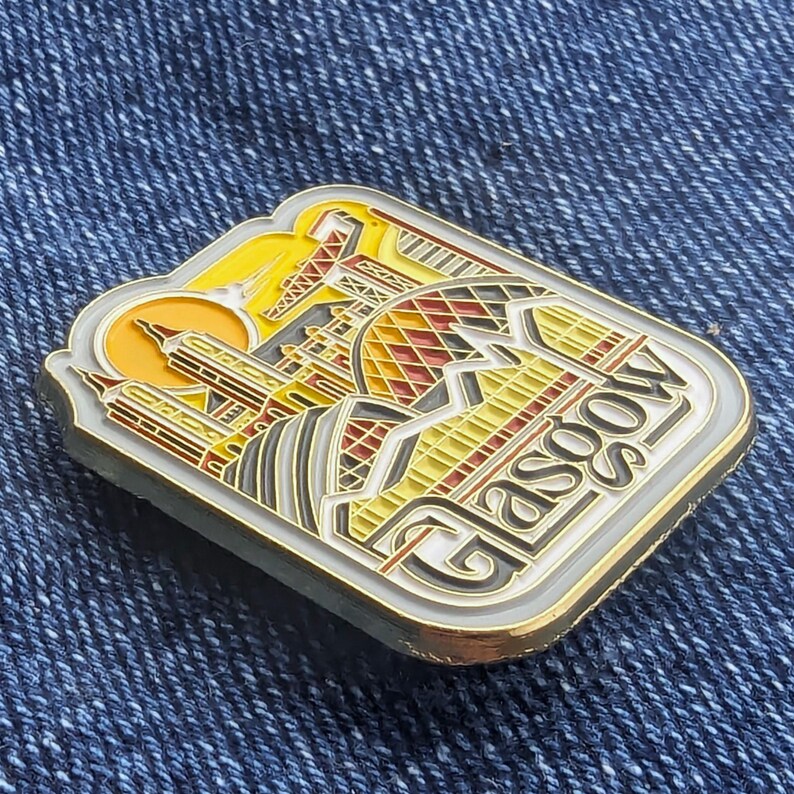 Peut inclure: &Eacute;pinglette rectangulaire repr&eacute;sentant un paysage urbain stylis&eacute; de Glasgow, avec le mot "Glasgow" en police d&eacute;corative. La broche a une bordure dor&eacute;e et un design aux tons jaunes, rouges et gris, sur fond de denim bleu.