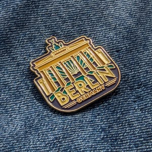 May include: Gold-colored enamel pin featuring a stylized depiction of the Brandenburg Gate with the word "BERLIN" and "GERMANY" below it. The pin has a blue, green, and yellow color scheme.