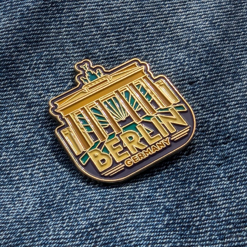 May include: Gold-colored enamel pin featuring a stylized depiction of the Brandenburg Gate with the word "BERLIN" and "GERMANY" below it. The pin has a blue, green, and yellow color scheme.