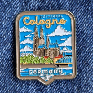 May include: A rectangular pin badge depicting Cologne Cathedral in Germany. The badge has a blue sky background with white clouds and the words "Cologne" and "Germany". The cathedral is brown and grey.