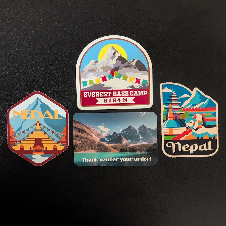 May include: Four colorful stickers featuring images of Nepal. The top sticker shows a mountain range with a yellow sun and the text "Everest Base Camp 5364 M". The sticker below it shows a mountain range with a lake and the text "Thank you for your order!" The sticker on the left shows a blue and red temple with the text "Nepal". The sticker on the right shows a colorful temple with the text "Nepal".