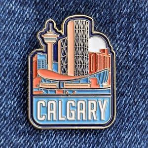 May include: A detailed enamel pin depicting the Calgary skyline, with buildings in orange, blue, and pink. The word "CALGARY" is displayed in white lettering at the bottom, framed by a navy blue border.