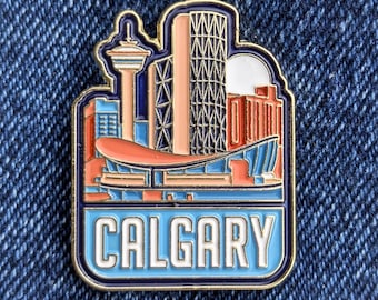 May include: A colorful enamel pin featuring a cityscape design with the word "CALGARY" in white lettering. The pin showcases iconic buildings in shades of orange, blue, and pink, outlined in gold and navy blue.