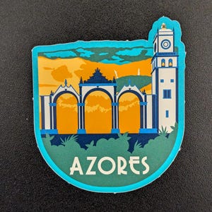 May include: A sticker featuring a scenic illustration of the Azores. The design includes a white and blue tower, arched structures, and the word "AZORES" in white. The background shows a sunset with orange and yellow hues.