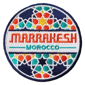May include: Circular embroidered patch. The design features the word "MARRAKESH" in orange, above "MOROCCO" in green. The patch has a white border and a navy blue background with geometric patterns in orange, teal, and yellow.