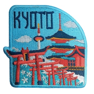May include: An embroidered patch featuring a light blue background with the text KYOTO in dark blue letters. The design depicts a red pagoda, a tall tower, and a series of red torii gates arranged in a row, accented with white cherry blossom motifs.