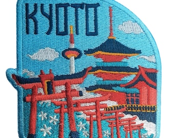 May include: An embroidered patch featuring a light blue background with the text KYOTO in dark blue letters. The design depicts a red pagoda, a tall tower, and a series of red torii gates arranged in a row, accented with white cherry blossom motifs.