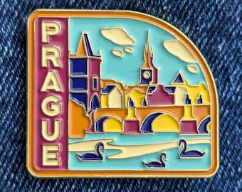 May include: A colorful enamel pin featuring a stylized cityscape of Prague. The pin has a gold border and includes the word "PRAGUE" in vertical letters. The design showcases buildings, bridges, and swans on a teal background.
