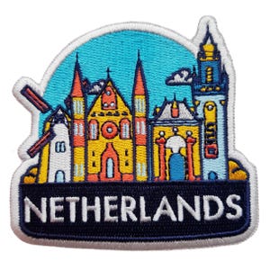 Puede incluir: Parche bordado con una colorida ilustración de arquitectura holandesa, incluyendo molinos y edificios, sobre un cielo azul. La palabra "NETHERLANDS" se muestra en texto blanco sobre una bandera azul marino.