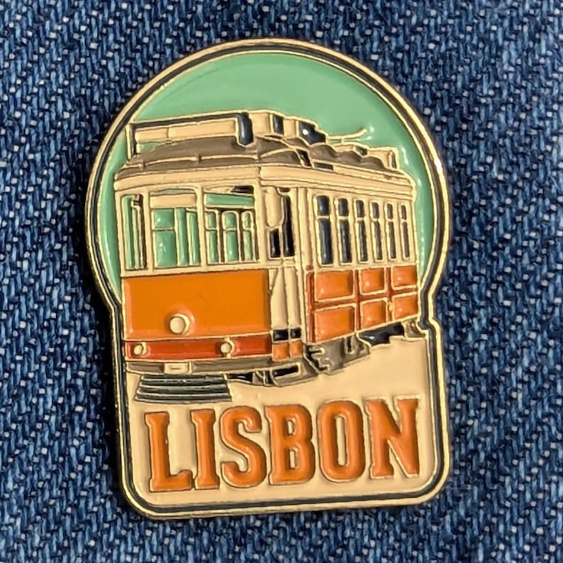 May include: A colorful enamel pin featuring a vintage orange and white trolley car with the word "LISBON" in orange letters. The pin has a gold border and a light green background.