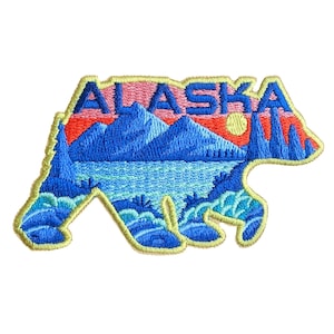 May include: Embroidered patch in the shape of a bear, featuring the word "ALASKA" in blue letters. The design includes a blue mountain range, a lake, and a sunset in shades of red and yellow. The patch has a yellow border.