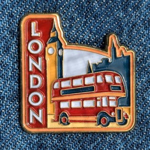 Könnte beinhalten: Ein rechteckiger London-Pin mit rotem Rand und dem Wort "LONDON" in Weiß. Der Pin zeigt einen roten Doppeldeckerbus, Big Ben und andere Londoner Wahrzeichen. Der Pin ist vor einem blauen Jeans-Hintergrund platziert.