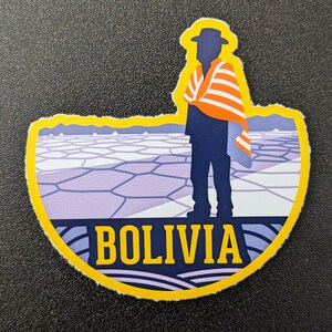 May include: A sticker featuring a silhouetted figure in a hat and poncho, standing before a landscape. The sticker has a yellow border and the word "BOLIVIA" in blue letters.
