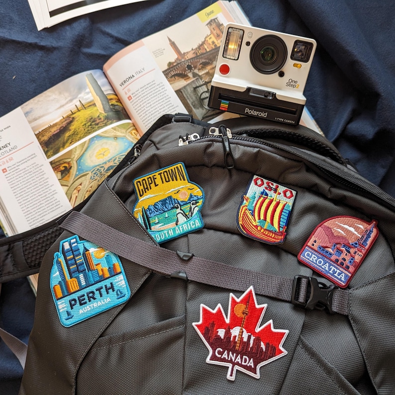 May include: A black backpack with travel patches for Cape Town, Oslo, Croatia, Perth, and Canada. A white Polaroid camera and a travel guide are also visible. The patches feature colorful illustrations of landmarks and cityscapes.