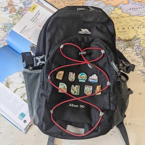 May include: Black backpack with red accents and travel pins. The backpack has a mesh side pocket and a red bungee cord design. A travel guide and a map are in the background.