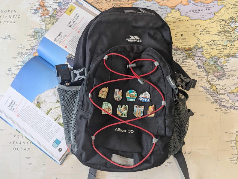 May include: Black backpack with red accents and travel pins. The backpack has a mesh side pocket and a red bungee cord design. A travel guide and a map are in the background.