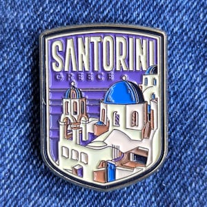 以下が含まれることがあります： ギリシャのサントリーニを描いた長方形のピン。ピンは、白い建物、青いドーム、およびテキスト「SANTORINI GREECE」を備えた紫色の背景を持っています。ピンは銀色で縁取られ、青いデニムの背景に配置されています。