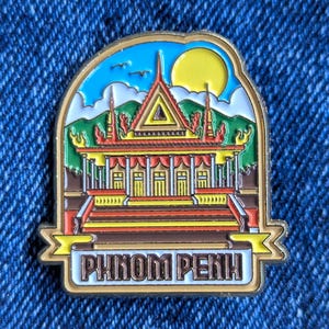 Pode incluir: Um alfinete dourado com uma ilustração colorida de um templo com o texto "PHNOM PENH". O alfinete tem um céu azul com um sol amarelo e montanhas verdes no fundo. O templo é vermelho, amarelo e castanho.
