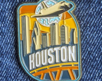 May include: A colourful enamel pin featuring a cityscape with the word "HOUSTON" in white lettering. The pin includes a space shuttle, a bridge, and a skyline in shades of orange, yellow, and blue.