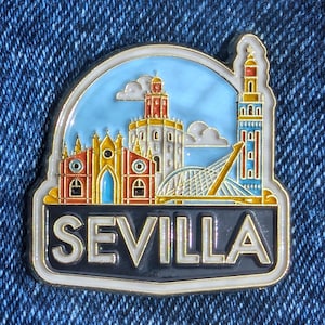 Könnte beinhalten: Ein farbenfroher Emaille-Pin mit einer Stadtlandschaft von Sevilla, Spanien, mit dem Wort "SEVILLA" in fetten Buchstaben. Der Pin zeigt ikonische Gebäude in Rot, Gelb und Blau vor einem hellblauen Himmel mit Wolken, umrahmt von einem goldenen Rand.