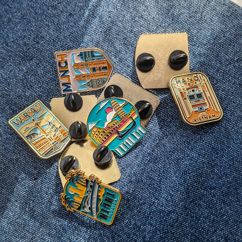 Op de afbeelding: Een verzameling emaille pins met reisbestemmingen. De pins tonen bezienswaardigheden en stadsnamen, waaronder "Warschau", "M&uuml;nchen", "Itali&euml;", "Hanoi" en "Miami". Elke pin heeft een uniek ontwerp en kleurenschema, met zwarte pin-backs.
