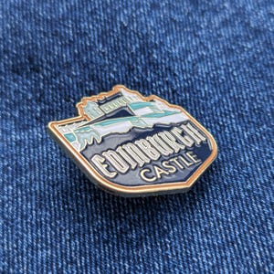 May include: An enamel pin depicting Edinburgh Castle, with a navy blue backdrop and white and teal details. The pin has a gold-coloured border and the words "EDINBURGH CASTLE" are written on it.