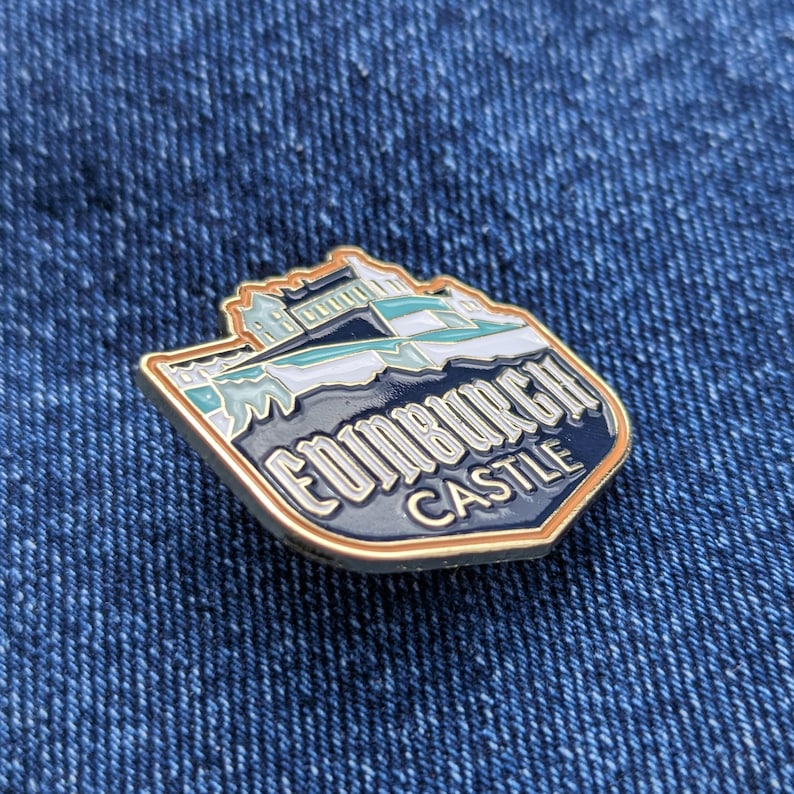 May include: An enamel pin depicting Edinburgh Castle, with a navy blue backdrop and white and teal details. The pin has a gold-coloured border and the words "EDINBURGH CASTLE" are written on it.