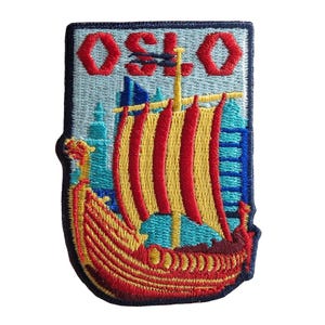 Puede incluir: Parche bordado que representa un barco vikingo con velas rojas y amarillas, sobre un fondo azul claro con edificios de la ciudad. La palabra "OSLO" está bordada en rojo en la parte superior. El parche tiene un borde azul oscuro.