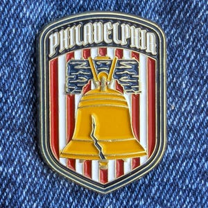 Puede incluir: Un pin con temática de Filadelfia con forma de escudo. El pin presenta la palabra "PHILADELPHIA" en la parte superior, con una Campana de la Libertad dorada en el centro. El fondo tiene rayas rojas y blancas, con detalles en azul.