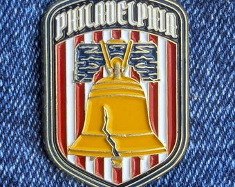 May include: A Philadelphia-themed pin in a shield shape. The pin features the word "PHILADELPHIA" at the top, with a gold Liberty Bell in the centre. The background has red and white stripes, with blue accents.