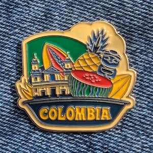 May include: A colorful enamel pin featuring a Colombian theme. The pin showcases a church, a pineapple, a watermelon, and a tiki-like figure. The word "COLOMBIA" is displayed in gold lettering on a dark blue background.