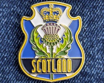 May include: A shield-shaped enamel pin featuring the Scottish flag design. The pin has a blue background with a white cross, a gold crown, and a thistle. The word "SCOTLAND" is written in gold.