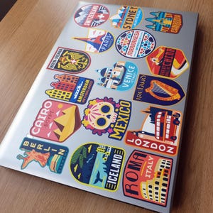 May include: A laptop covered in colorful travel stickers featuring cityscapes, landmarks, and country names. The stickers include destinations like Berlin, Cairo, Paris, Sydney, Venice, and London.