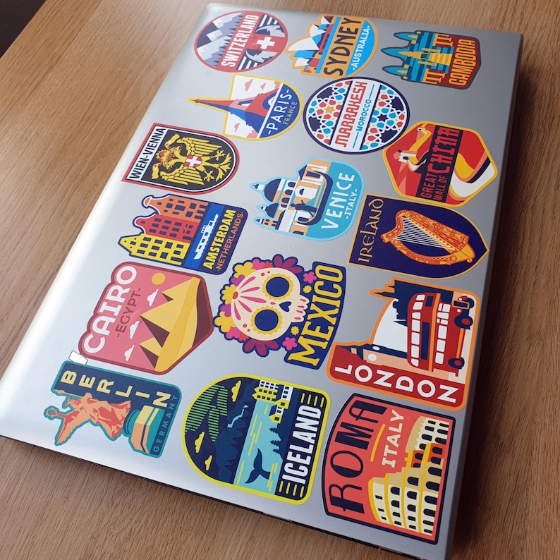 May include: A laptop covered in colorful travel stickers featuring cityscapes, landmarks, and country names. The stickers include destinations like Berlin, Cairo, Paris, Sydney, Venice, and London.