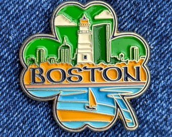 May include: A clover-shaped enamel pin with a Boston theme. The pin features a lighthouse, skyline, and sailboat over a blue and orange water scene. The word "BOSTON" is displayed in orange letters.